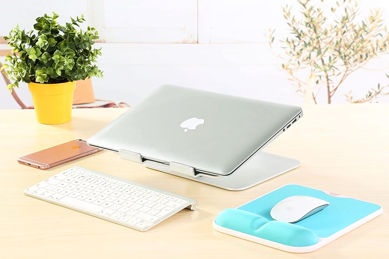 A desk with a silver aluminium laptop stand with an apple macbook placed on it. The laptop is closed.  Also on the  desk is a plant in a yellow pot, an apple iphone, a white wireless keyboard, and a white wireless mouse on a blue mouse mat. 