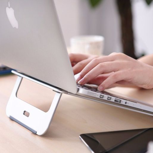 A MacBook laptop, elevated on very thin but strong looking laptop stand made from aluminium. A person's hands are typing 