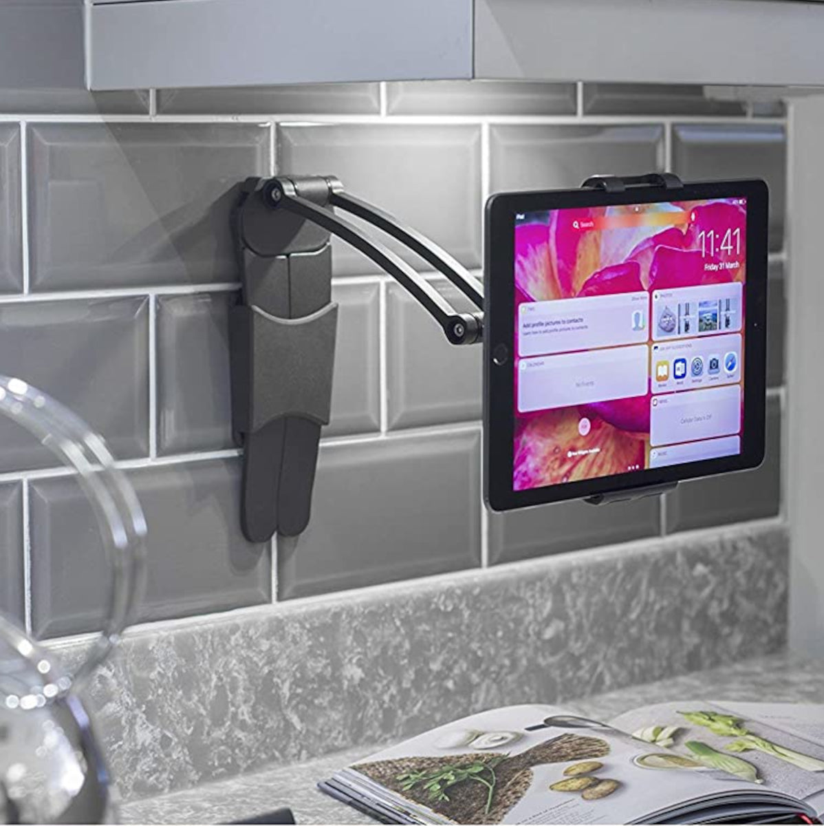  A kitchen with an Apple Ipad Pro held on a wall mounted tablet stand.  The 5mm alen bolts which lock the angles can be seen on the side of the stand in 2 places.  The tablet is around 30cm from the wall. The wall has grey subway tiles with white grout.  A recipe book showing parsley, mushrooms, new potatoes and some greens is open on the kitchen counter 